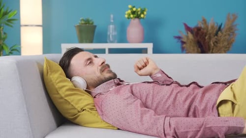Man relaxing on sofa at home with headphones listening to music, daydreaming and happy.