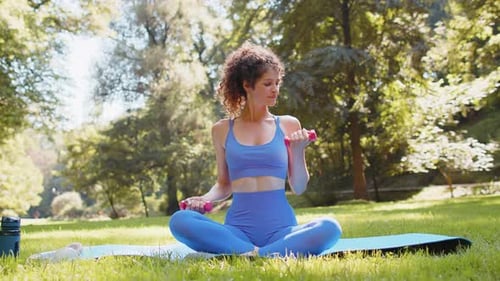Sportive Woman Exercising Weightlifting with Dumbbells Sitting on Yoga Mat in Park on Sunny Day