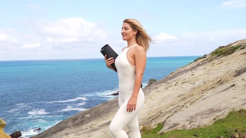 Blonde woman holding yoga mat smiles by the ocean