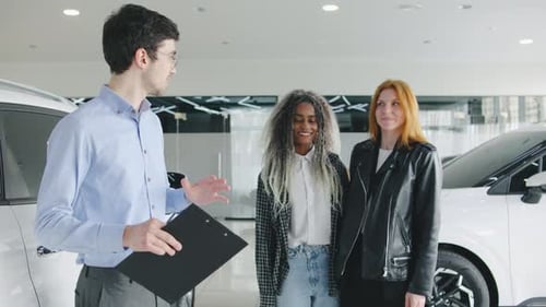 A Lesbian Couple Tours a Modern Car Dealership with a Male Salesman Listening Attentively As He