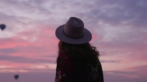 Young Girl Admiring The Hot Air Balloons Flying In Cappadocia At Sunrise In Turkey. - rear view