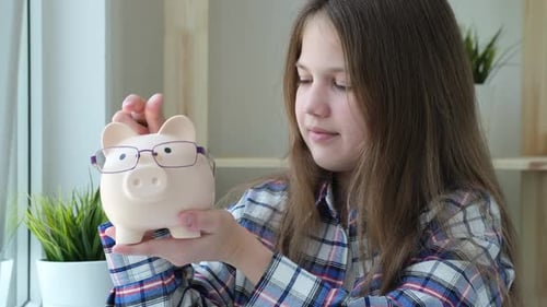 Smiling Preteen Girl Holding Pink Piggy Bank in Glasses and Putting Coins in It
