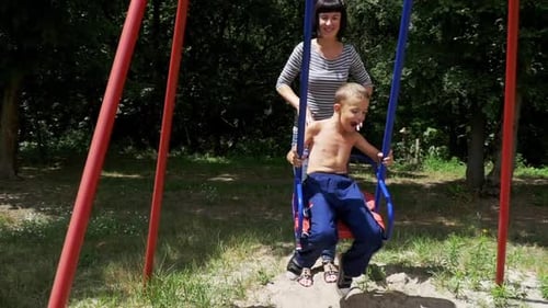 Mother Swinging Her Happy Child Boy on a Street Swing at Playground in Slow Motion