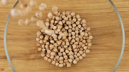 Chickpea seeds are poured into a glass bowl against a wooden oak board background. Top view. Slowmo