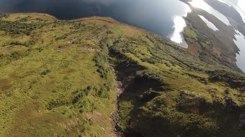 Flying along a mountain overlooking a lake and grassy landscape and many trees on the hillside.