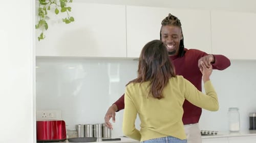 Casal diversificado estendendo as mãos, girando debaixo do braço, se abraçando e sorrindo na cozinha ao lado de uma torradeira vermelha