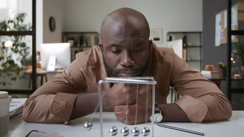 Portrait of Man Looking at Camera from Behind Newtons Cradle on his Office Desk