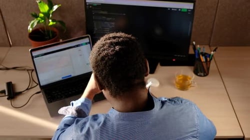 Homem negro escrevendo linhas de código em um PC de mesa com dois monitores e um laptop ao lado em um escritório elegante