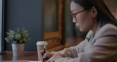 Young Asian Lady Writing in Notebook and Working with Laptop Computer Focused on Business Planning