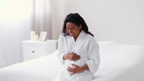 Frau in weißer Robe sitzt auf dem Bett und hat während der Schwangerschaft Beschwerden