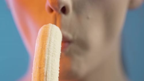 Close Up Of Woman's Mouth Eating Banana Fruit, Studio Shot With Colorful Lighting