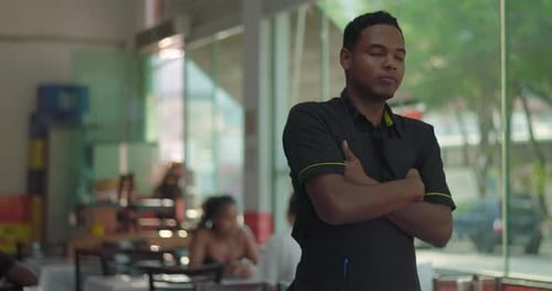 Camarero masculino confiado con los brazos cruzados posando seriamente cerca de la ventana del restaurante, comedor interior