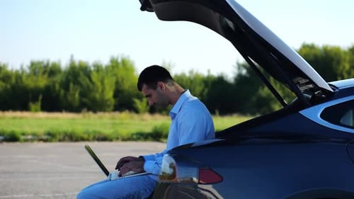 Young Handsome Entrepreneur Works on Notebook Sitting in Electric Car Trunk Successful Businessman