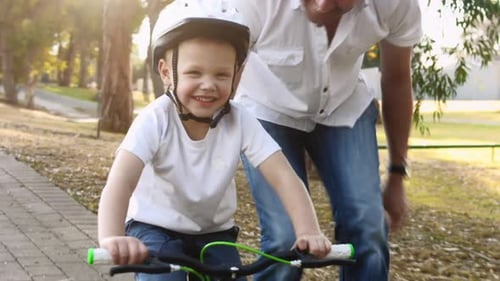 Father teaches young son to ride bike on park pathway with safety helmet
