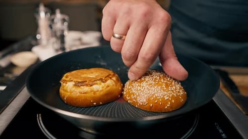 A close-up of a chef toasting a bun on a griddle while cooking a burger in a restaurant kitchen