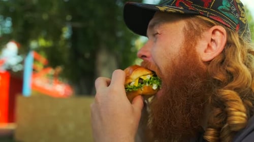 Bearded hungry man eating an appetizing burger sitting in the cafe outdoors