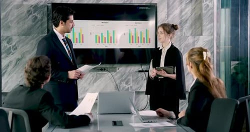 Asian businessman leading professional meeting while discussing bar chart data on screen with female