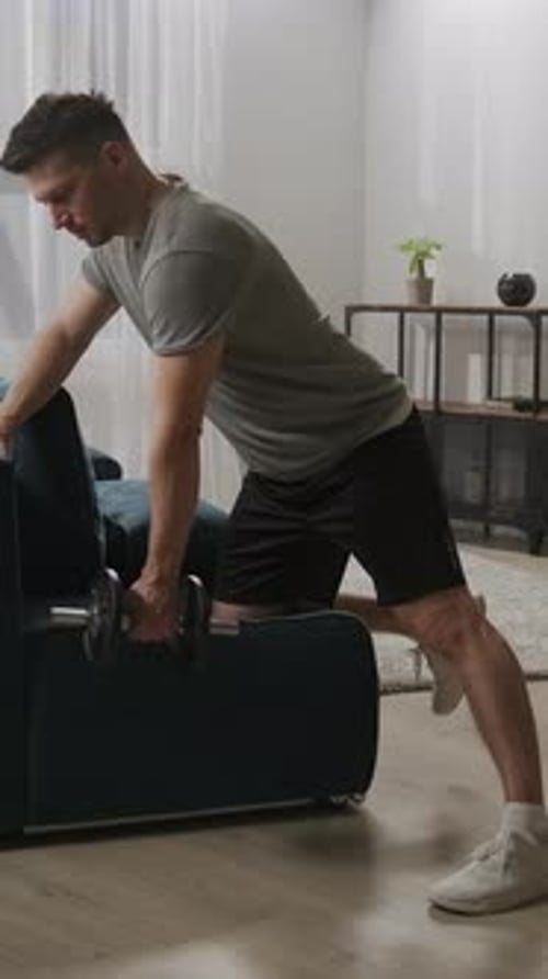 Focused Home Workout with a Man Performing Dumbbell Rows Using a Sofa for Support in a Bright Living