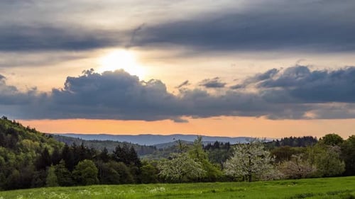 Sonniger Sonnenuntergang an bewölktem Himmel erhellt ein hügeliges Tal mit blühenden Obstgärten, Wiesen und Wäldern
