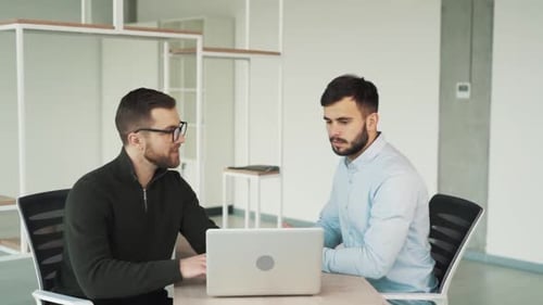 Two young men are discussing work while sitting in the office at the laptop