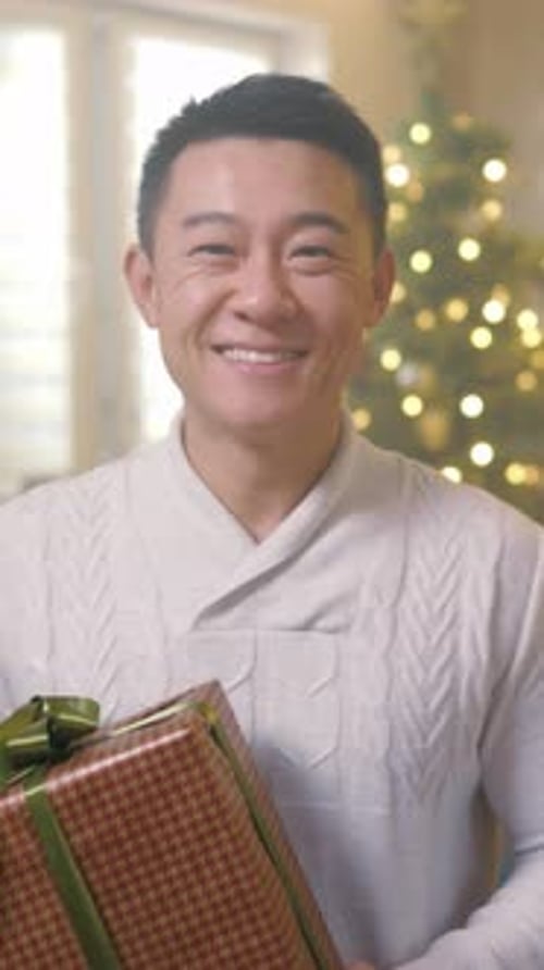 Smiling man holds Christmas gift indoors by tree