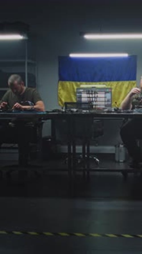 Worker in Camouflage Uniform Moves Equipment Cases Through Drone Facility While Team Works on Drone