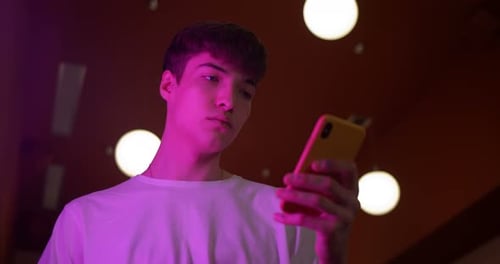 Young Millenial Guy Wearing White TShirt Using His Smartphone Standing at Futuristic Neon Lights