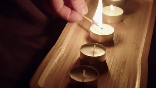 New tea candles are lit with wooden match flame in dark room tray
