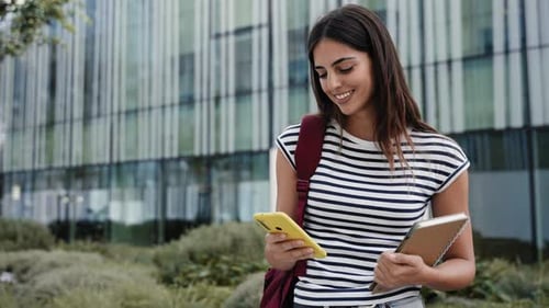Happy Hispanic Student Girl Using Yellow Cell Phone Outdoors at University Campus Smiling Young