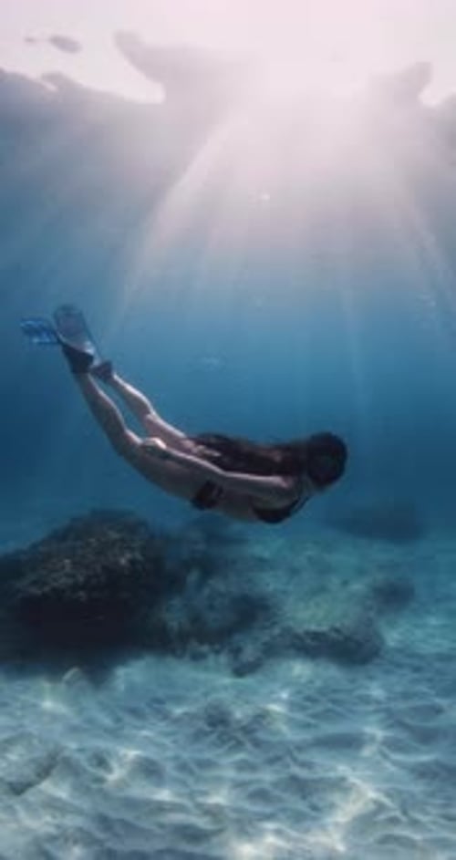 Freediver Woman with Freedive Fins Swims Underwater in Blue Sea with Undersea Sun Rays