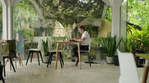 Une professionnelle transgenre travaille assidûment sur un ordinateur portable dans un espace de coworking moderne entouré d'une végétation luxuriante