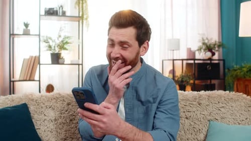 Man Laughing While Reading Funny Social Media Comments on Smartphone Relaxing on Comfortable Sofa