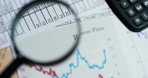 A hand with a magnifying glass passes over a financial newspaper and financial bag with a business