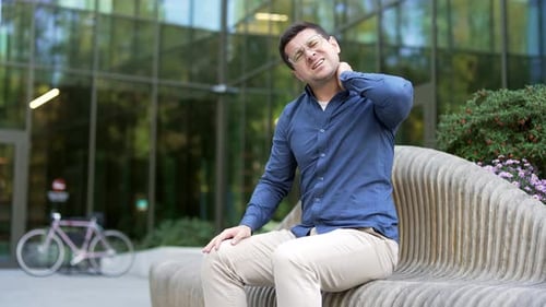 Tired businessman suffering from neck pain while sitting on a bench on the street near a modern