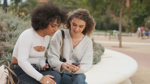 Two Happy Friends Using the Mobile While Sitting on a Public Park