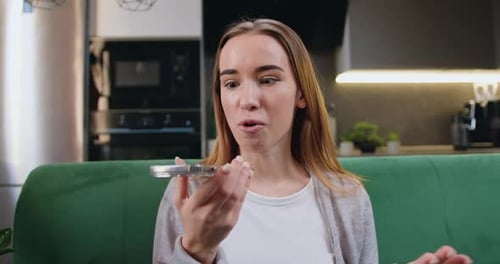 Hermosa chica alegre que usa el teléfono inteligente para grabar mensajes de voz mientras está sentada en un acogedor sofá