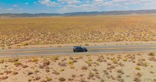 Cars going by the empty road in the sunny desert. Mountainous horizon line under blue cloudy sky