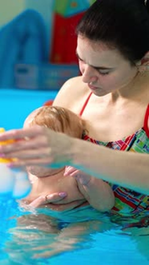 Woman is holding a cute little infant in the swimming pool. Coach is pouring the kid from a toy.