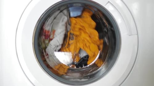 Rotierende Waschmaschinentrommel, bei der bunte, nasse Kleidungsstücke fallen, während sie Wasser und Reinigungsmittel spritzen