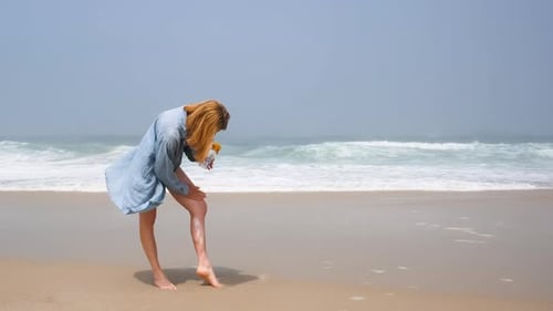 Young Woman Applying Sunscreen to Her Leg While Standing on a Sunny Beach Dressed in Casual