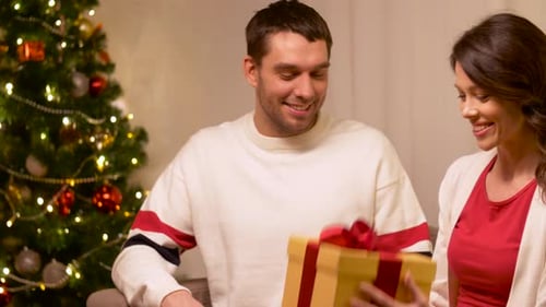 Woman Gives Christmas Present To Man by Christmas Tree