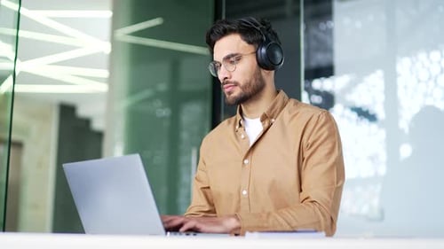 Confident IT specialist developer in wireless headphones works on laptop computer sitting
