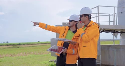 Zwei kaukasische Ingenieure mit orangefarbener Sicherheitsjacke und Helm diskutieren über das Windturbinensystem