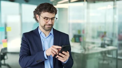 Confident businessman in a suit is using phone standing at workplace in business office. Man worker