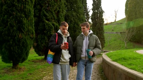 Young caucasian gay couple walking in park, holding hands and looking at smartphone
