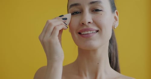 Woman Applying Makeup with Sponge on Yellow Background