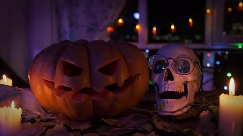 Close up, carved pumpkin glowing inside next to skull on dry foliage near colorful garlands