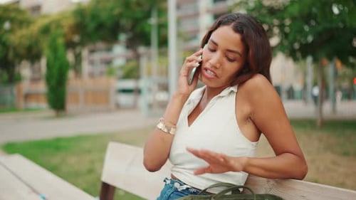Girl with long dark hair wearing white top sits on bench in city park and uses mobile phone
