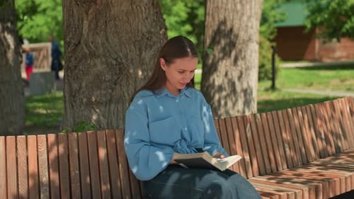 Young White Woman Reading on Bench in Sunlit Park Under Oak Tree Denim Shirt Turning Pages with Calm