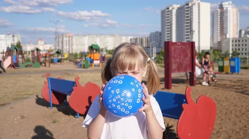Girl Inflates a Blue Balloon on the Playground in Summer